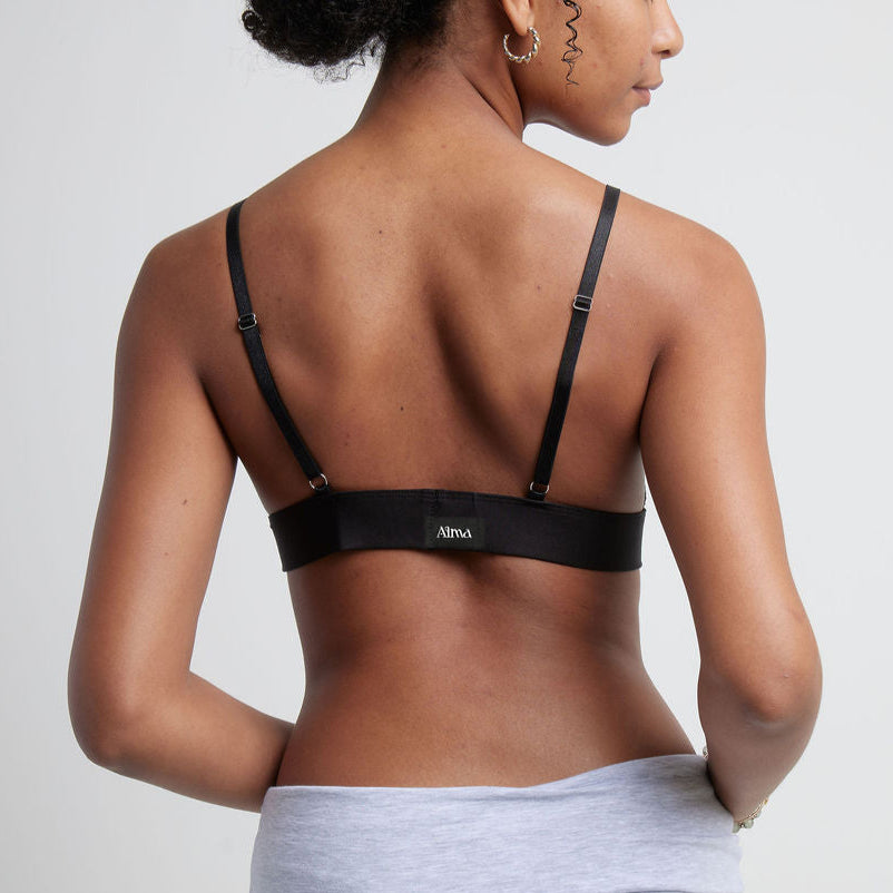 A woman wearing a black Alma bralette, with her back to the camera, against a neutral wall.