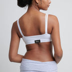 Woman wearing a white Alma bralette, facing with her back to the camera, against a neutral wall.