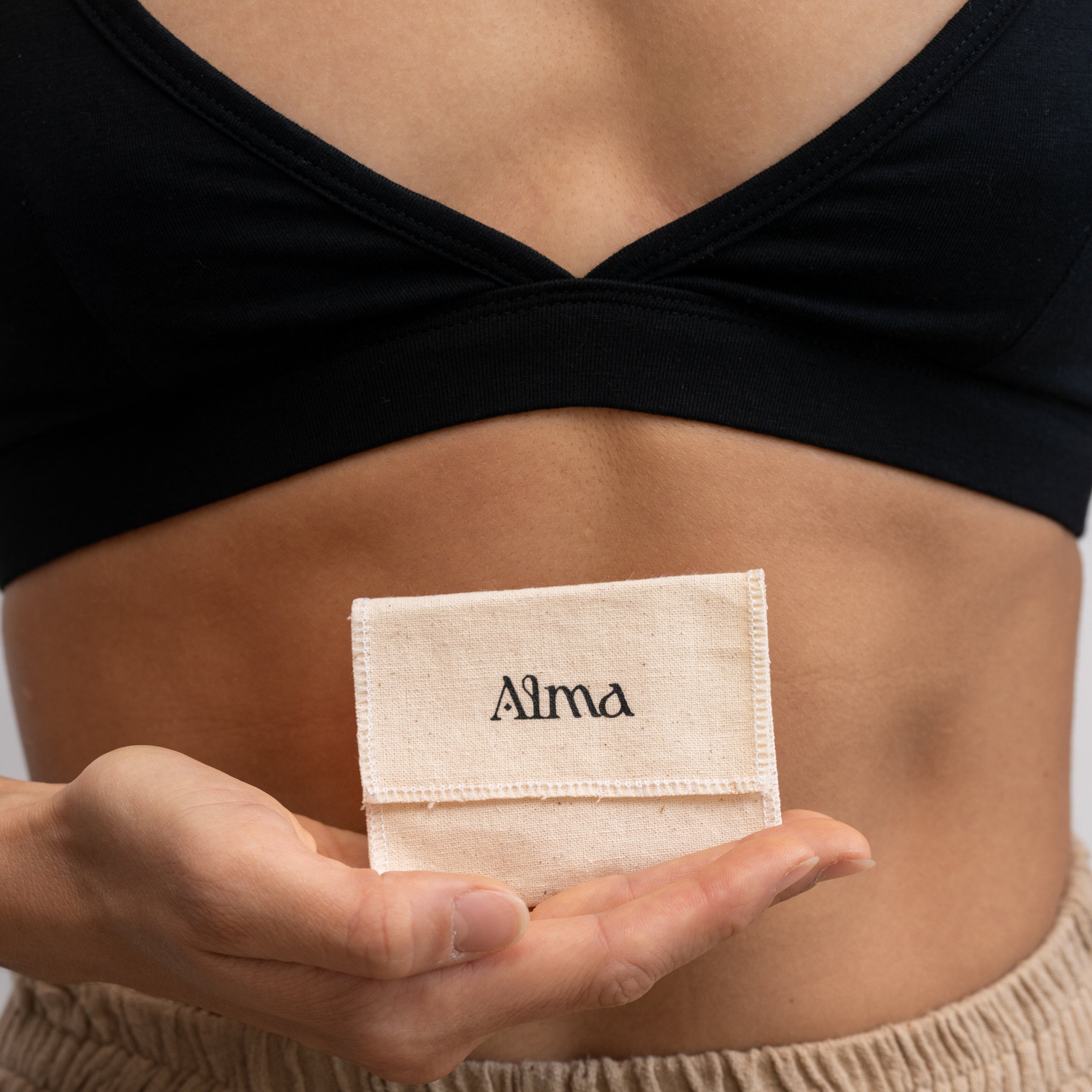 A woman wearing a black Alma bralette with neutral-coloured pants, holding a small fabric pouch with 'Alma' branding on the front.