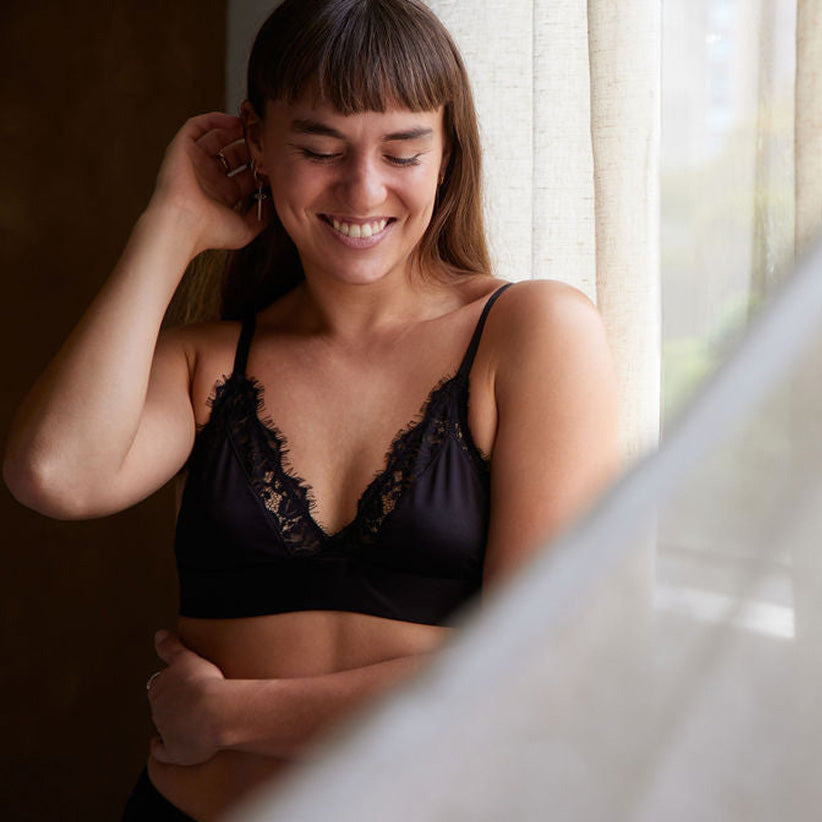A woman wearing an Alma Avara bralette, standing against a window, in a softly lit room.